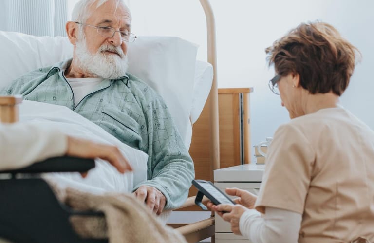 Caregiver kneeling down beside hospital bed asking an elderly man some questions